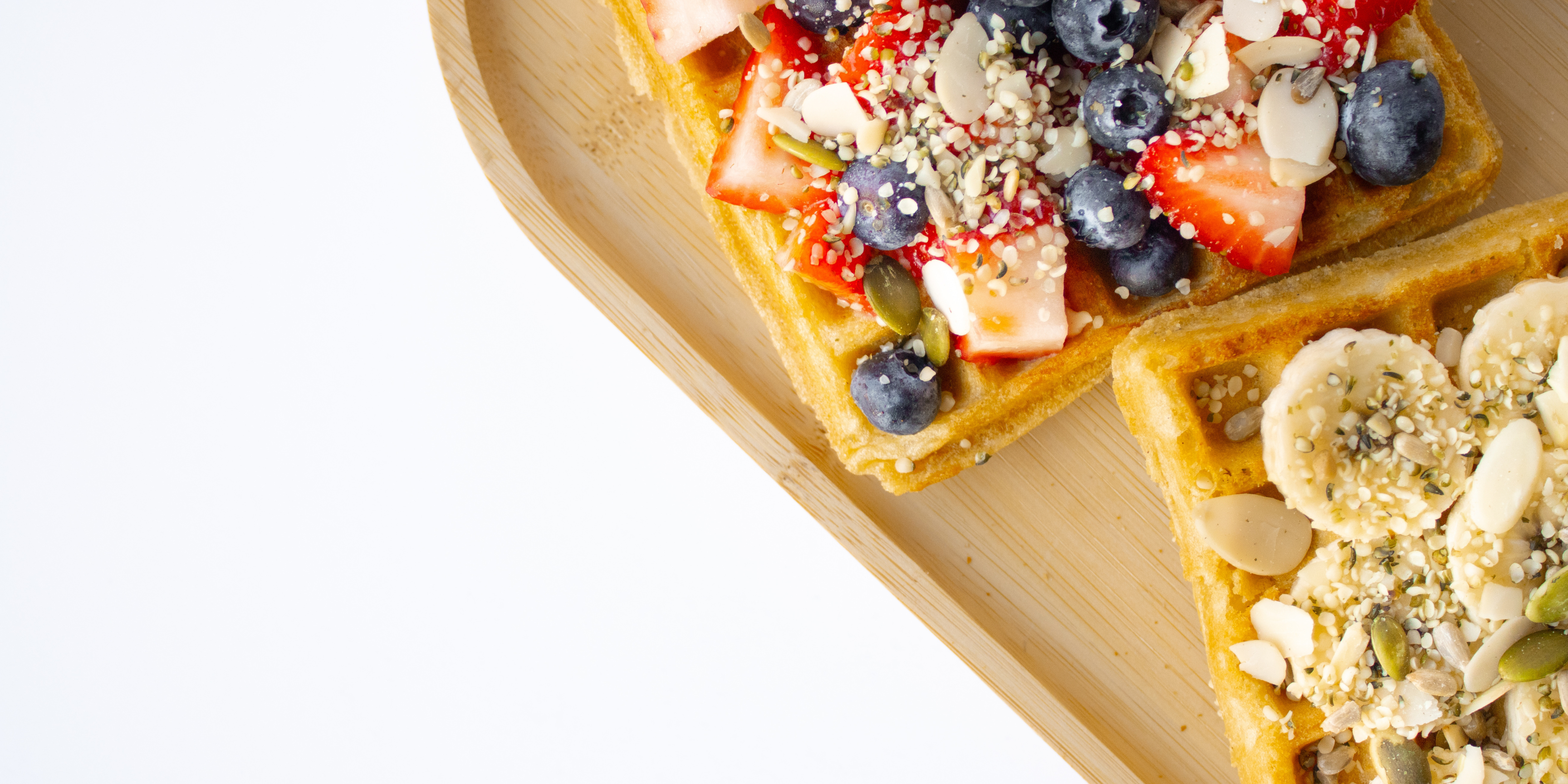 berry waffles and banana waffles on a bamboo plate covered in nuts and seeds against a white backdrop