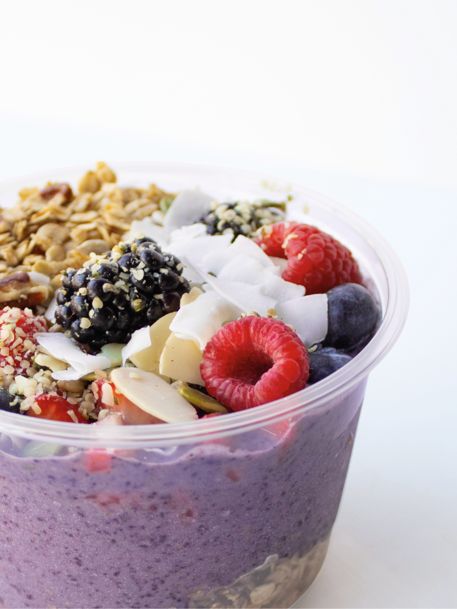 purple acai bowl with granola, raspberries, blueberries, blackberries, coconut shavings, and seeds against a white backdrop