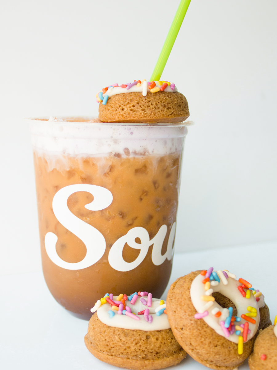 ube latte with purple cold foam with a green straw, surrounded by mini sprinkle donuts against a white backdrop