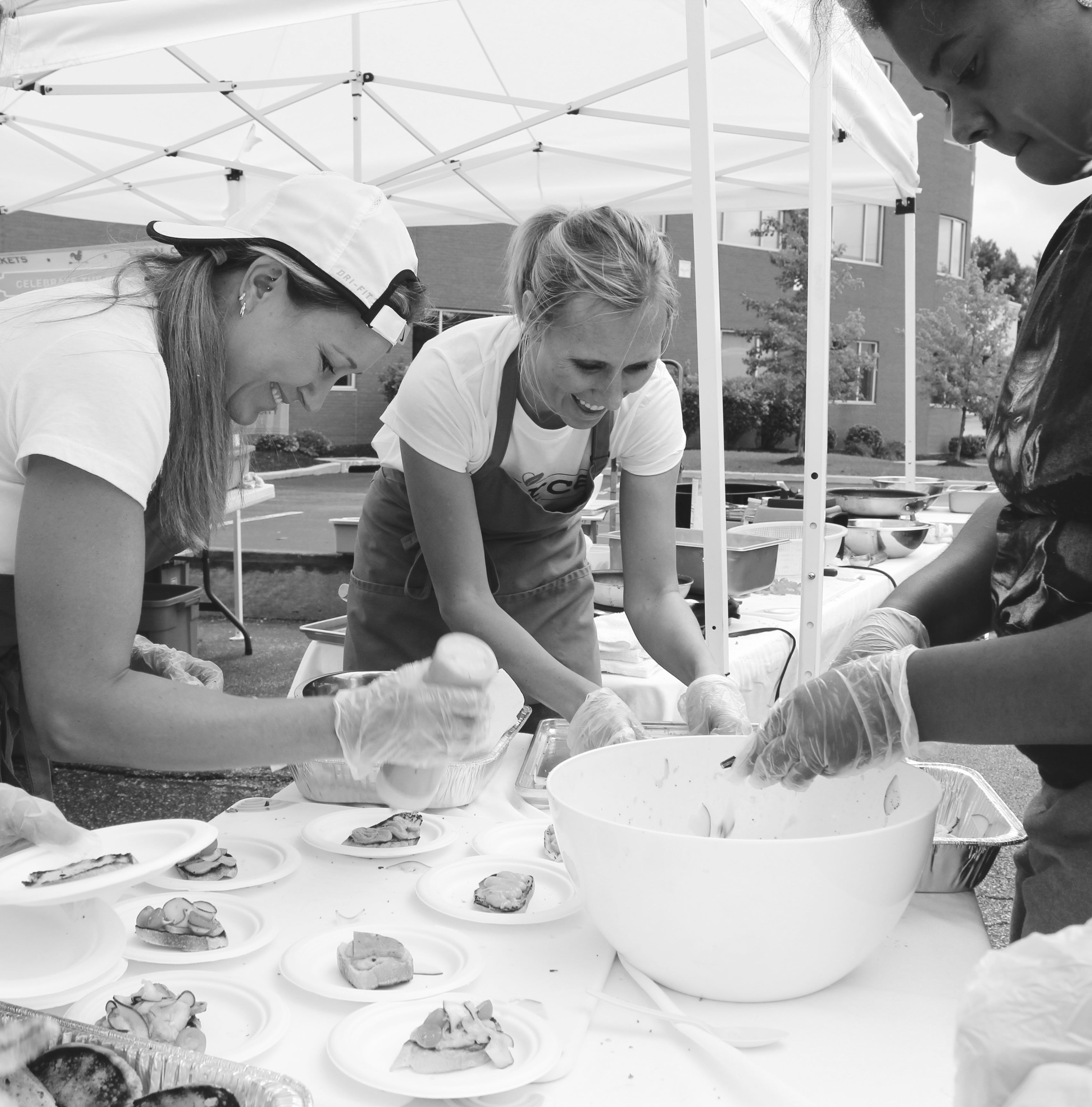 three women serving small plates of food in a tent outside while laughing together