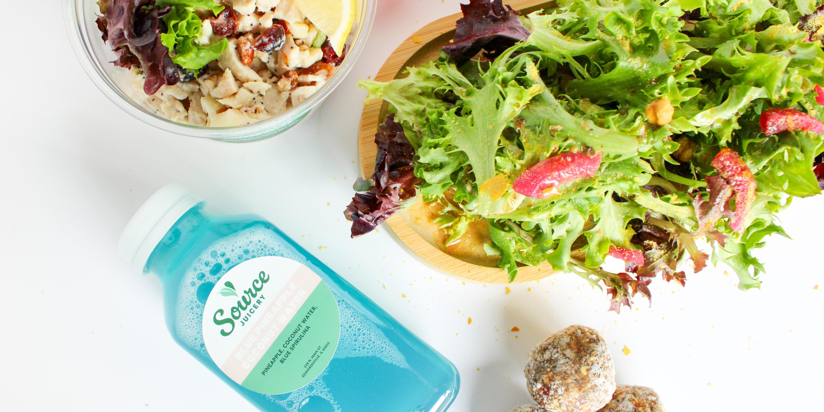 Overhead shot of a blue pineapple water, bright green salad with red onions, house made chicken salad and protein power bites from Source Juicery in Edwardsville, IL