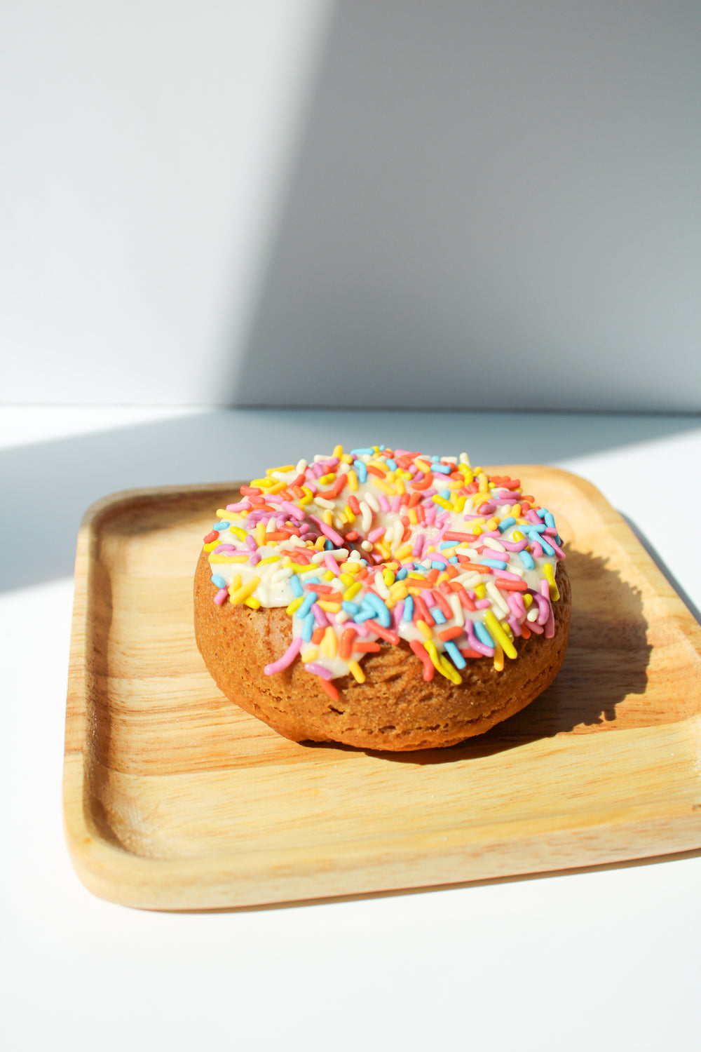 gluten free vegan vanilla donut with white glaze and rainbow sprinkles on a bamboo plate against a white background
