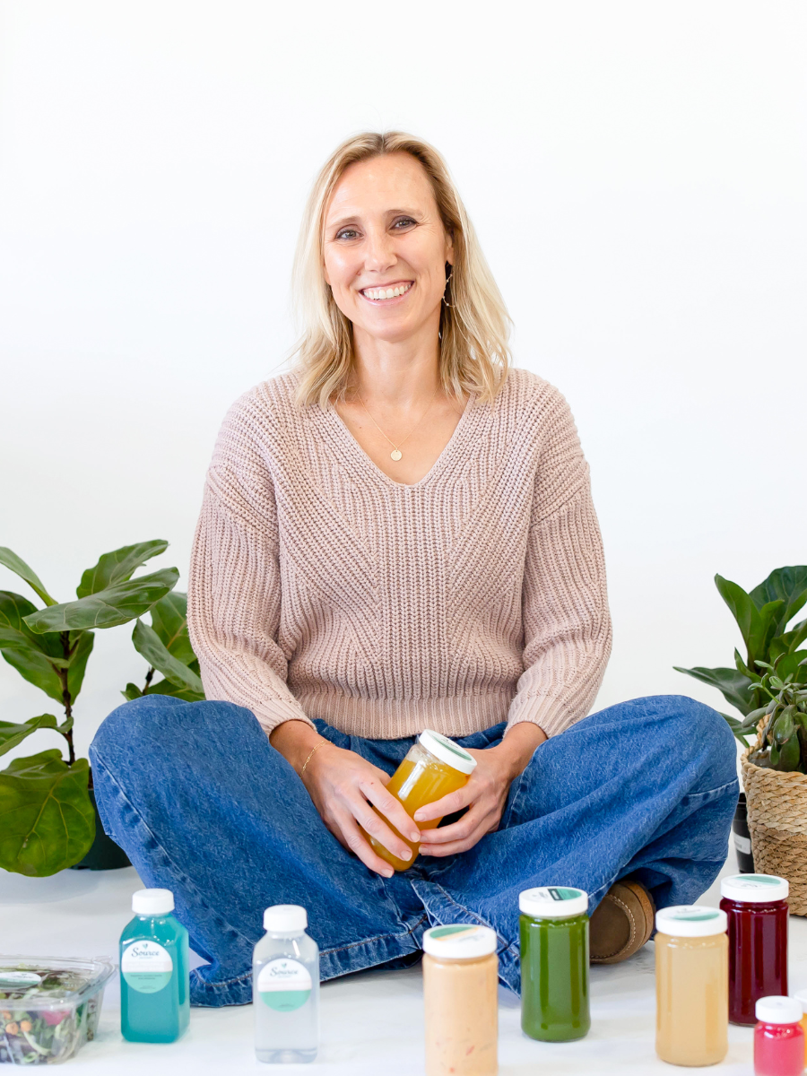 blonde woman in a beige sweater and jeans sitting on the ground cross legged with juice and plants against a white backdrop
